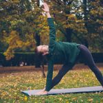 Cheerful young woman is doing yoga balancing exercises during individual practice in city park standing on mat and enjoying rest and nature. People, healthy lifestyle and sports concept.